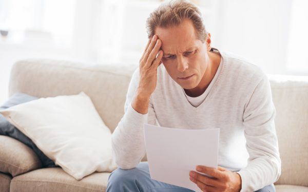 Depressed mature man holding paper and looking at it while sitting on the couch at home